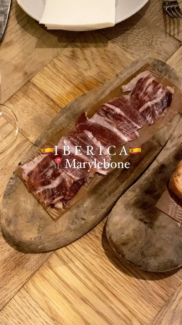 The video showcases a meal at Iberica restaurant in Marylebone.  It features various dishes, including cured ham, pan con tomate, patatas bravas, meatballs with mashed potato, black rice with prawns, and churros with chocolate sauce. The food is presented on wooden boards and in simple, elegant bowls. The video also includes a shot of the restaurant interior and a wine bottle.