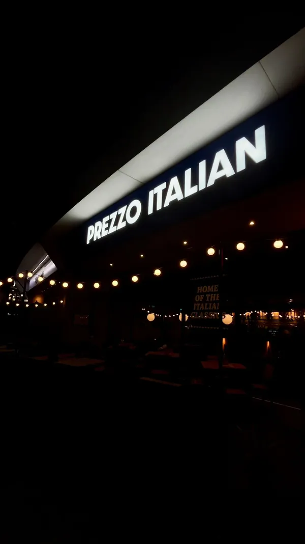 The video showcases a dining experience at Prezzo, an Italian restaurant in Chelmsford. It begins with exterior shots of the restaurant at night, highlighting its signage. The video then moves inside, showing the cozy, modern interior, a cocktail, and a gluten-free menu. Several dishes are featured in close-up shots, including a burrata and tomato salad with a 'Gluten-Free' flag, a bowl of olives, a creamy chicken and mushroom fusilli pasta, a baked lobster mac and cheese, and truffle fries. The video concludes by showing the dessert menu and a slice of chocolate fudge cake with vanilla ice cream, which has a celebratory sparkler in it.
