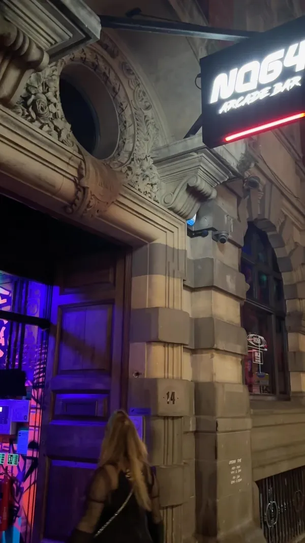 The video showcases NQ64, a retro arcade bar in Bristol. It features various arcade games, including Pac-Man and a Simpsons game, as well as console games like FIFA. The bar offers a selection of cocktails; the video highlights the 'Kong Island Iced Tea', 'Hits From the Pong', and 'Kirby' cocktails, emphasizing the Halloween-themed drinks and a color-changing ice cube. The overall atmosphere is vibrant and energetic, with Halloween decorations visible throughout the bar.