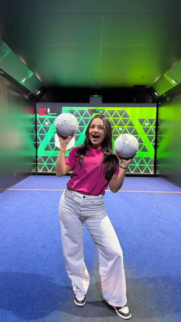 A woman showcases her experience at Toca Social, an interactive football and entertainment venue. The video features clips of her and her friends playing various virtual football games, enjoying cocktails, and eating food. The food shown includes buffalo chicken wings, mini cheeseburger sliders, and fries. The venue has a modern, dark aesthetic with neon lighting and large screens for both the games and watching live sports.
