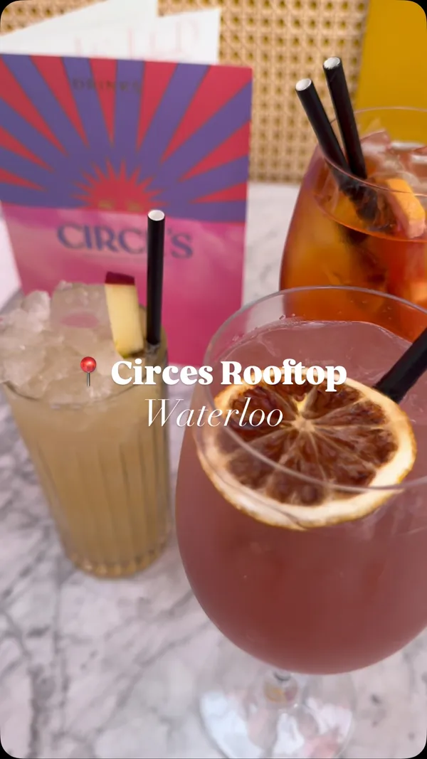 The video showcases a bustling rooftop bar called Circe's. It features shots of the venue's decor, including large straw light fixtures and plants, and patrons socializing. The video highlights cocktails, showing a bartender preparing a red drink (likely an Aperol Spritz) and presenting three different cocktails on a marble table: a red one, a pink one garnished with a dried orange slice, and a lighter coloured one with crushed ice and an apple slice garnish. A menu for Circe's is also visible. The video briefly shows a woman drawing.