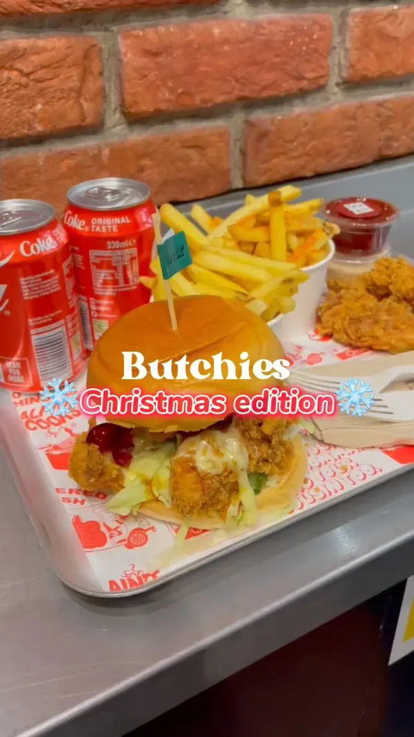 This video showcases the 'Sweet Baby Cheesus' Christmas burger at Butchies, a restaurant with various locations. The video features close-ups of the burger, highlighting its crispy chicken tenders, camembert cheese, cranberry sauce, and chilli honey.  It also shows crispy chicken tenders and spicy fries as sides, alongside Coca-Cola.  The setting is primarily the restaurant itself, with shots of the food and merchandise.