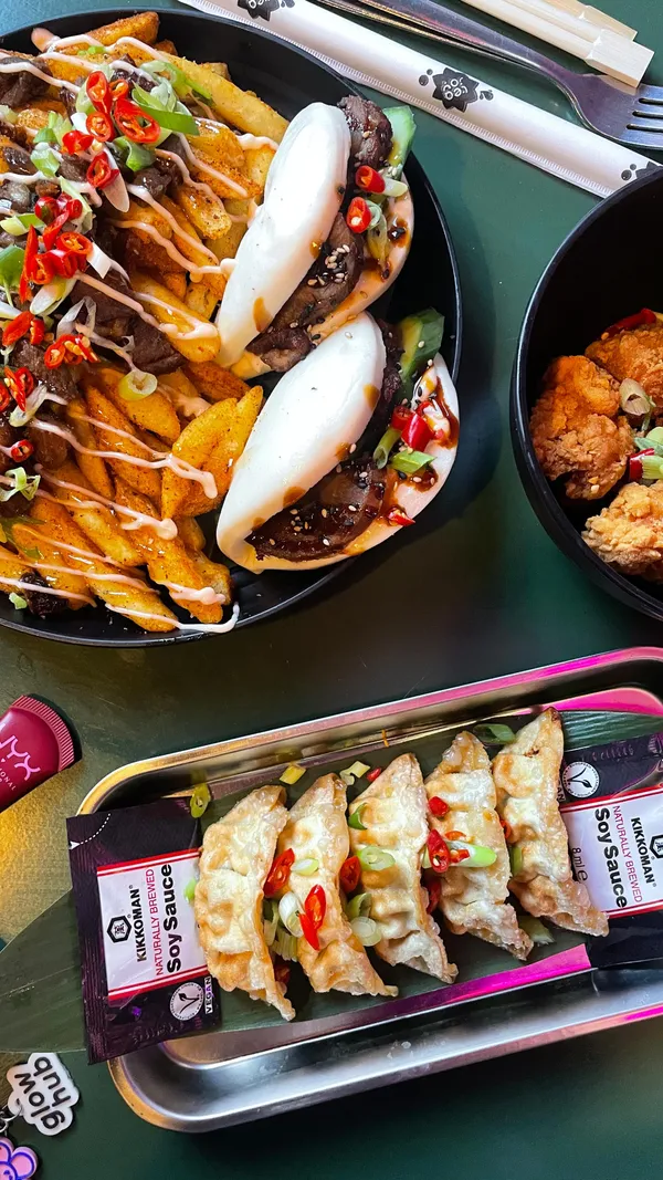 The video showcases a visit to Inamo Sukoshi, a Japanese fusion food vendor inside the Cutlery Works food hall in Sheffield. It begins with two women outside the venue, then shows the vendor's stall. The main focus is on the food, featuring close-up shots of several dishes: fried chicken karaage, a platter with two duck bao buns and loaded duck fries, and chicken gyoza. The video also shows one of the women eating the gyoza and chicken karaage with chopsticks. A mango bubble tea is also featured. The video ends with a shot of a Christmas tree inside the venue.