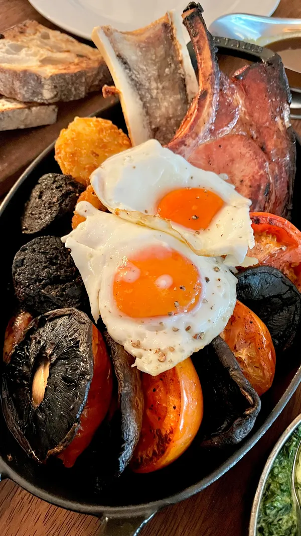 This video showcases a luxurious Hawksmoor restaurant breakfast experience for two. The setting is a high-end restaurant, elegantly decorated. The food includes a full English breakfast platter with a unique addition of a sugar pit bacon chop and grilled bone marrow, alongside fried eggs, grilled mushrooms and tomatoes, and baked beans.  A fillet carpaccio, Cornflake Bourbon milkshake, and sticky toffee pudding are also presented. The drinks showcased are an espresso martini and Irish coffee. The food is meticulously presented in various dishes, highlighting the ingredients’ quality and the restaurant’s attention to detail. No specific dietary information is provided.