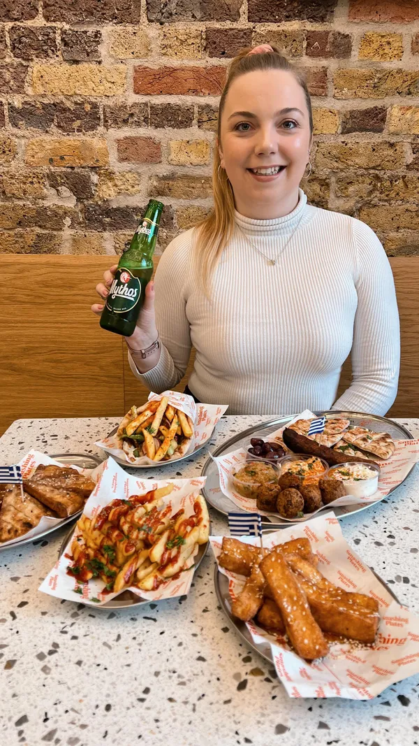 The video showcases a meal at Smashing Plates, a Greek restaurant. It features various dishes: a meze platter (tzatziki, smoked aubergine, dips, pita, olives, pork sausage, falafel); chicken gyros; spicy fries; and halloumi fries with syrup and sesame seeds.  The food is presented on metal trays with branded paper. The setting is the restaurant itself. The video's primary focus is on showcasing the food and drinks.