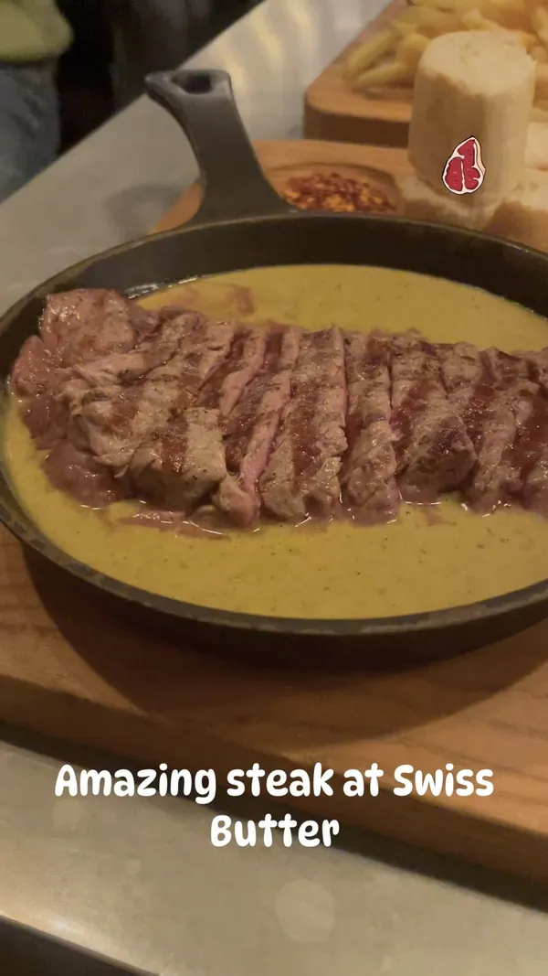 The video showcases a meal at Swiss Butter restaurant in Holborn. The main focus is a sliced steak in a creamy green sauce, served with french fries, sliced baguette, and a side salad. The presentation is visually appealing, with the food arranged attractively on a wooden platter.  The video doesn't explicitly mention any dietary information.