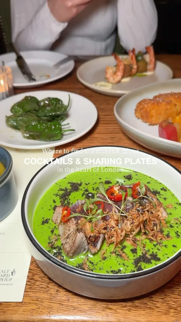 The video showcases a meal at Ember Yard restaurant in London. It features various sharing plates and cocktails, including prawns with prawn croquetas, yellowfin tuna with guindilla ajoblanco, grilled Ibérico with romesco, Rubia Gallega pinchos, heirloom tomato salad with fontina cheese, grilled asparagus, and coffee cheesecake. The cocktails shown include a pink margarita-style drink and a whiskey-based cocktail. The food is presented beautifully, with careful plating and close-up shots highlighting the textures and colors of the dishes. The video focuses on a meal for two and highlights the restaurant's atmosphere and the quality of the food and drinks.