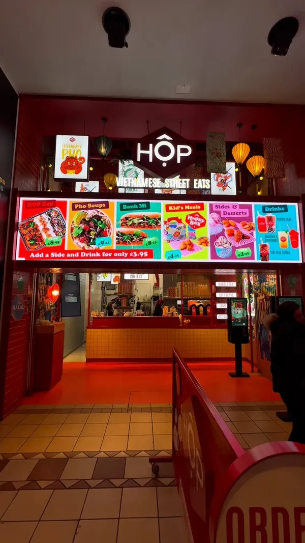 The video begins with an exterior shot of the HOP Vietnamese Street Eats restaurant entrance, showing the menu board. It then transitions to a table displaying a variety of takeaway food items, including containers of fried items (possibly tater tots or similar) with dipping sauces, spring rolls, a large sandwich (likely Banh Mi), a cup of Pho soup, a drink cup, and canned beverages. Close-up shots feature adding chili oil to the Pho, holding up a spring roll, and pouring sauce onto the sandwich.