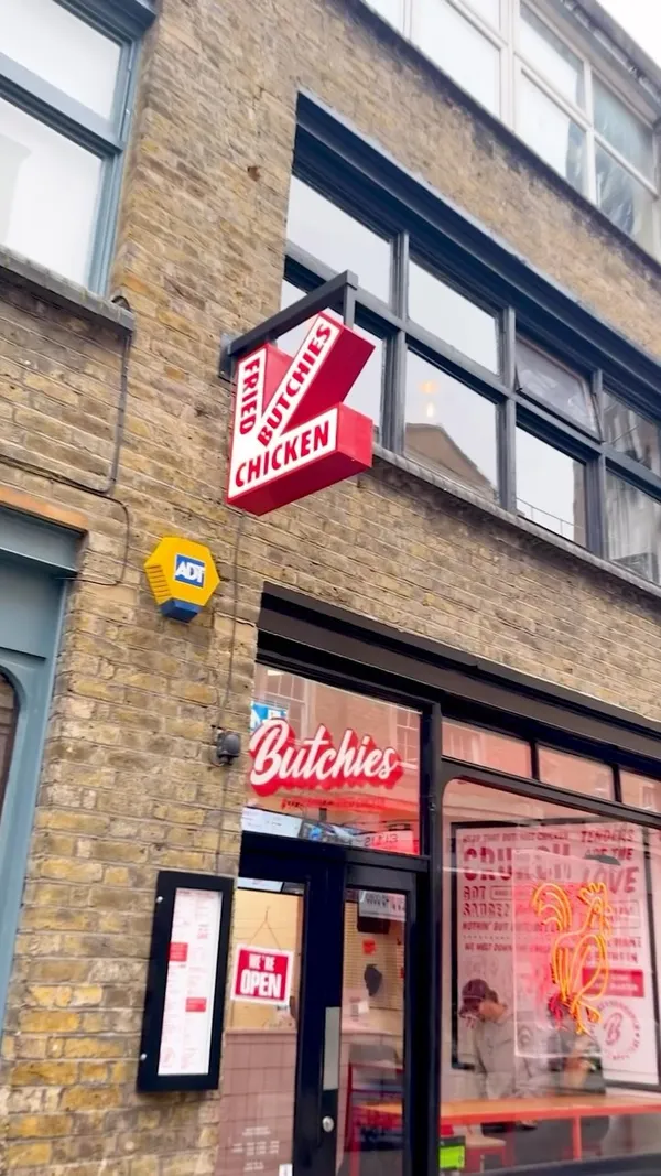 The video showcases Butchies Fried Chicken restaurant in Shoreditch, London. It features the restaurant's exterior and interior, highlighting the menu, which includes fried buttermilk chicken sandwiches (Buffalo, Cheesy Rider, The OG, BBQ, Jenny from the Block), chicken wings, popcorn chicken, and sides like fries and slaw.  Plant-based options are also shown. The video focuses on a meal deal, showing the preparation of the OG chicken burger and fried chicken tenders, which are then covered with gravy.  The meal is served with fries and Camden Hells Lager. Dipping sauces are also prominently displayed.