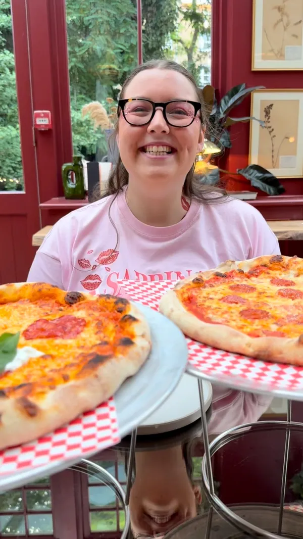 The video showcases a visit to a stylish restaurant, Eve Kensington. It features a woman happily receiving two large pizzas. There are multiple shots of the restaurant's interior, which has a conservatory-like feel with large windows, red-painted frames, checkered floors, lush plants, and eclectic, colourful furniture. The food shown includes two types of pizza (one pepperoni, one with salami and burrata), and a side of parmesan fries. Key food moments include a cheese pull, dipping the pizza crust into a sauce, and taking a bite. The overall vibe is fun, positive, and aesthetically pleasing.