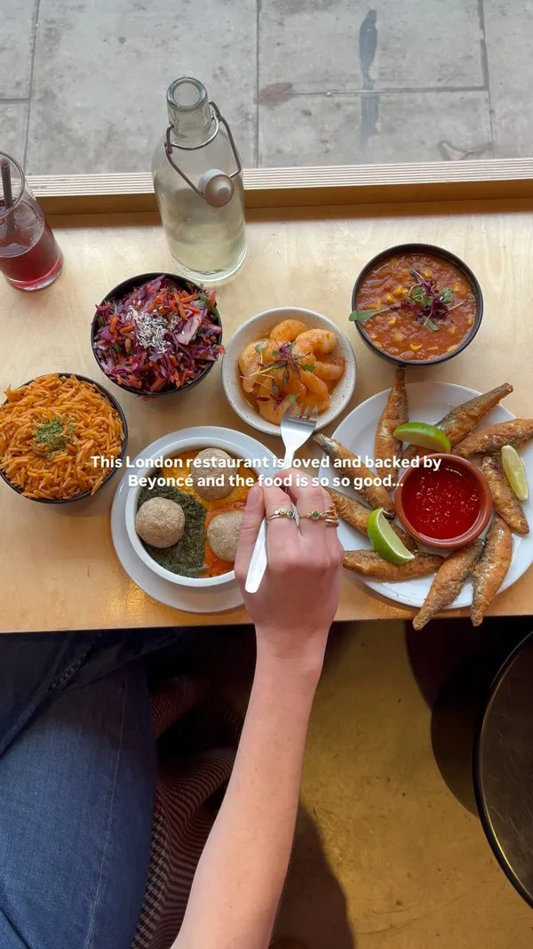 The video showcases a variety of Nigerian dishes served tapas-style at Chukus restaurant in North London. The setting is a table inside the restaurant, with views of the street outside. The food items shown include Jollof rice, a red cabbage salad, a dish with three different sauces and three balls, honey suya prawns, fried fish with lime and a red dipping sauce, and lime and ginger cheesecake. The presentation emphasizes the variety and colorful nature of the dishes. The main focus is on showcasing the restaurant's menu and highlighting its appeal, including its endorsement by Beyonce.
