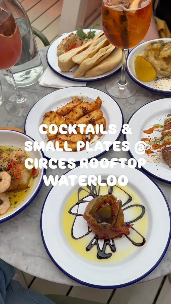 The video showcases a rooftop bar and restaurant experience. It features various small plates including hummus with pita bread, fried halloumi, roasted fig wrapped in prosciutto, spiced lamb kofta skewers, tiger prawns on toast, and tempura calamari. Several cocktails are also shown, including an Aperol Spritz-like drink, a pink cocktail garnished with a dried lemon slice, and a green cocktail. The video includes shots of the venue's interior, seating areas, and views. Hands are shown interacting with the food (dipping pita, cutting fig, cutting prawns) and drinks (toasting). A text overlay at the beginning identifies the venue and offering.