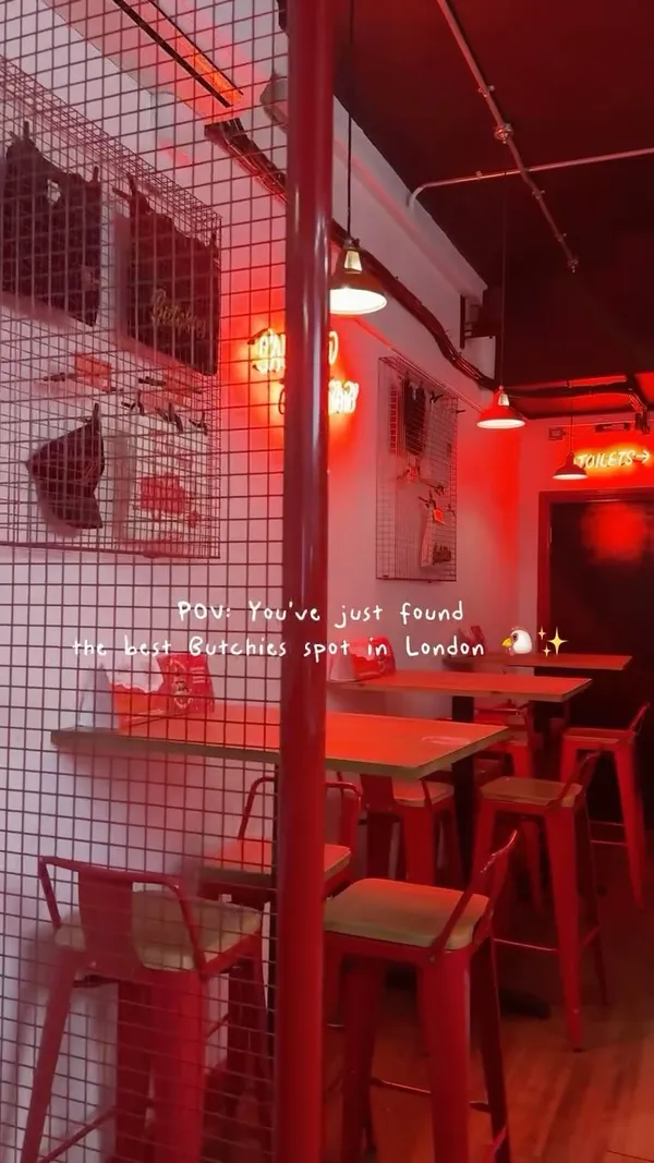 The video showcases Butchies, a fried chicken restaurant in London. It features a young woman enjoying a meal that includes a burger, fries, and chicken tenders.  The setting is the restaurant itself, decorated in red and white. The food is presented on trays; close-up shots highlight the textures of the fried chicken and the burger's contents. No specific dietary information is provided. The main focus is on the food and the enjoyable dining experience.