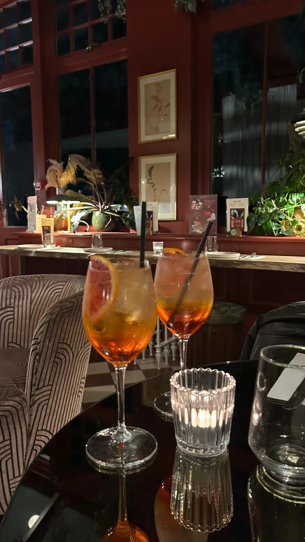 The video showcases the Eve Kensington restaurant, highlighting its pizza and cocktail offerings. The setting is a stylish restaurant with red accents and indoor plants. The video features Aperol Spritz cocktails with orange slices and black straws, followed by shots of two pizzas: one with pepperoni and mozzarella, and another with sausage and red onion. Buffalo fried chicken wings with a dipping sauce are also shown. The pizza bases are described as thin. The main focus is on showcasing the food and drinks available at the restaurant.