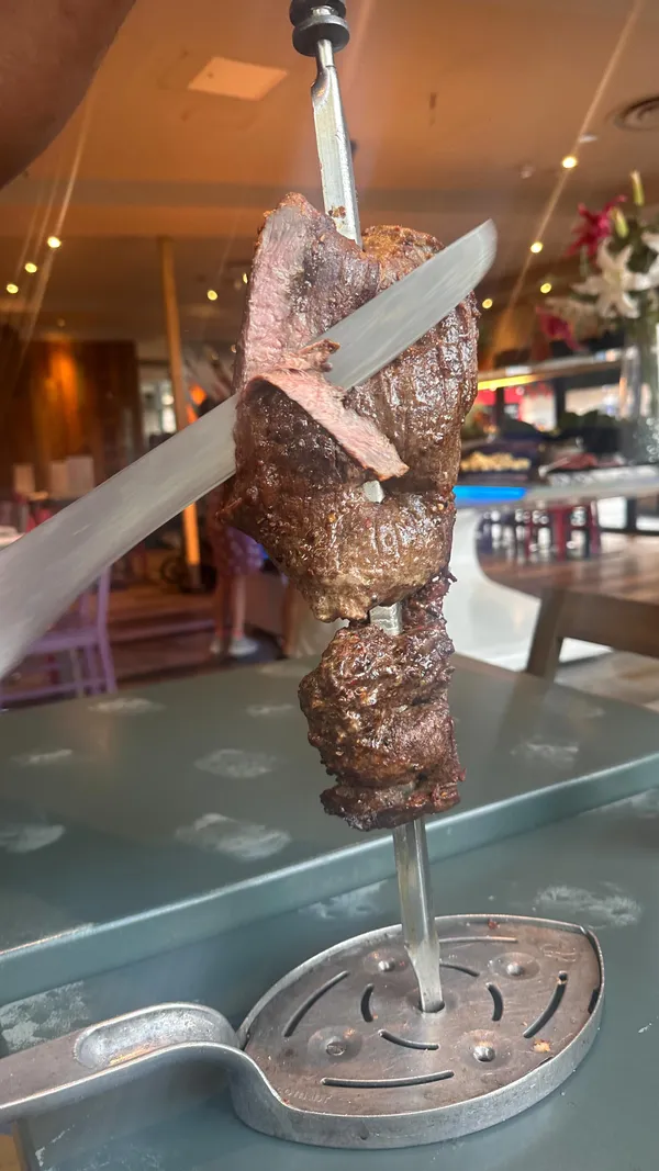 The video promotes Preto, a Brazilian steakhouse in London. It begins with an exterior shot of the restaurant, followed by a view of the hot buffet which includes items like caponata, french fries, and hot butternut squash. The video then shows the restaurant's system for requesting more meat using a 'More Please' card. The main focus is on the rodizio experience, with various meats being carved from large skewers at the table, including different cuts of steak, sausages, and chicken wings. A shot of grilled pineapple being carved is also featured. The video concludes by showcasing two desserts: a slice of cheesecake with caramel sauce and a chocolate pudding with ice cream and chocolate sprinkles.