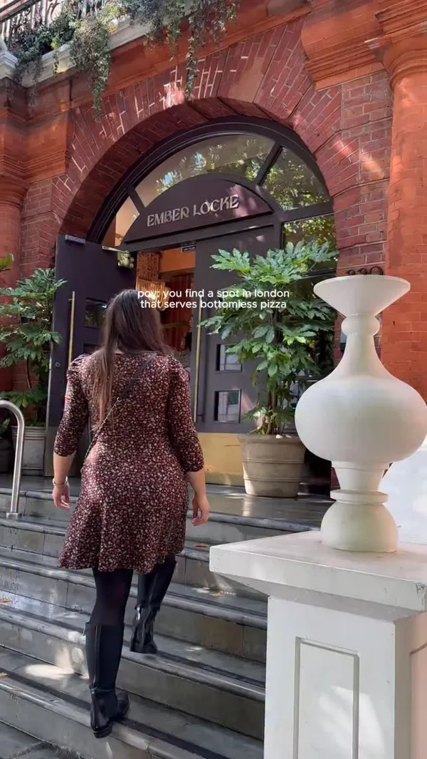 The video promotes a restaurant called Ember Locke in Earl's Court, London, focusing on its 'bottomless pizza' offer. It begins with a woman entering the establishment, followed by clips showcasing the stylish garden and indoor seating areas. The video features several appetizing shots of different pizzas, including a pepperoni pizza with a prominent cheese pull, and another pizza topped with a whole burrata being cut into. The overall vibe is trendy and appealing, highlighting both the food and the aesthetic of the venue.