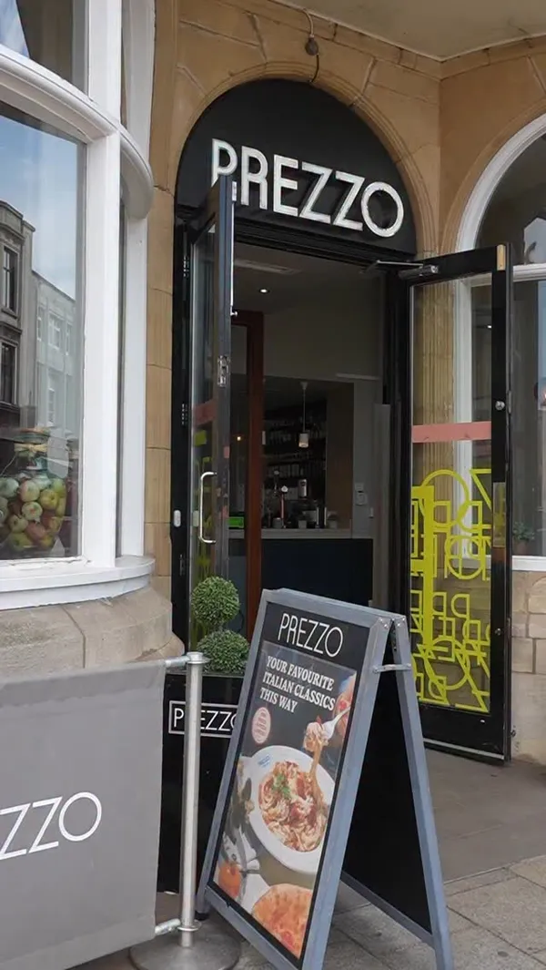 The video provides a tour of the Italian restaurant, Prezzo. It begins with an exterior shot of the restaurant's entrance, then moves to the al fresco dining patio. The video showcases two cocktails being toasted, followed by various dishes including grilled bread with olive oil and balsamic vinegar, a creamy ravioli dish with shrimp and tomatoes getting cheese grated on top, fried mozzarella sticks, a red sauce ravioli also getting cheese, and a pizza with chicken and sausage being sliced with a pizza cutter. The final shots show an overhead view of the table laden with food and another close-up of the cocktails.