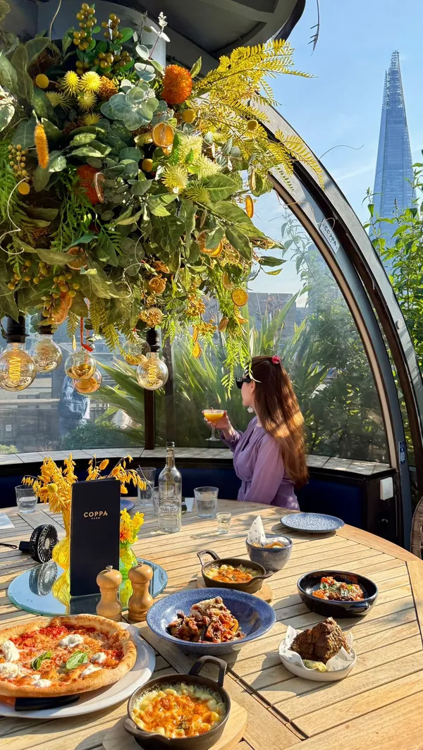 A video montage showcasing a lunch experience at Coppa Club by Tower Bridge in London on a sunny day. The video features shots of the restaurant's famous igloos and outdoor terrace, with iconic views of Tower Bridge and The Shard. Various dishes are presented, including a margherita pizza being sliced, a bowl of pappardelle pasta with ragu, mac and cheese, and other small plates. Desserts shown include a crème brûlée and a bowl of assorted ice cream, both enjoyed with the scenic backdrop.