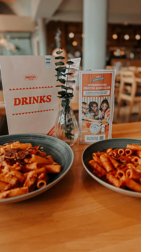 The video promotes the Italian restaurant chain Prezzo, specifically highlighting its new 'Kids Club'. It begins with an exterior shot of the restaurant, then moves inside to show the table setting and a young girl coloring with the kids' activity pack. The video showcases several dishes, including a chicken schnitzel with spaghetti and two different rigatoni pasta dishes. A key shot features parmesan cheese being freshly grated over the schnitzel dish. The video concludes with the young girl happily eating.