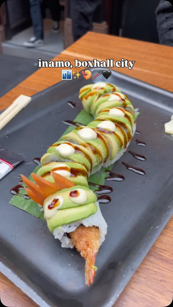 The video is a short vlog-style review of Inamo Sukoshi, a sushi restaurant located in Box Hall City, London. It begins with the creator and a friend outside the venue, then shows the interior of the food hall and the Inamo stall. The video showcases the menu, a lunch deal sign, and three different sushi rolls served on black trays. The featured dishes include a creatively presented Dragon Roll (tempura shrimp and avocado, shaped like a dragon), a Spicy Tuna Roll, and another roll topped with crispy onions. The creator is shown eating the sushi with chopsticks. The voiceover describes the venue as a perfect spot for a city lunch and praises the sushi for being fresh and well-portioned.