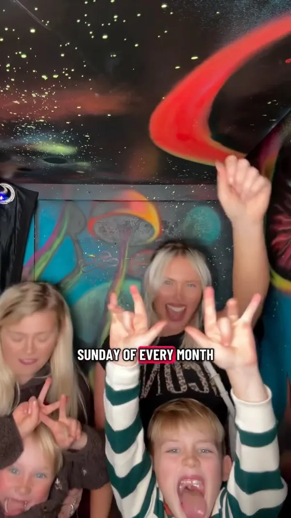 The video promotes NQ64, a retro arcade bar in Edinburgh, highlighting their family-friendly event on the last Sunday of each month. It follows a mother and her two young children as they enter the venue and enjoy the experience. The video showcases the bar's unique, dark atmosphere with neon and UV lighting. It features a bartender preparing drinks, including one with a dramatic flame effect and two colourful mocktails. The children are shown enthusiastically playing various arcade games, such as a driving simulator, a fighting game, a light gun shooter, and a guitar-based rhythm game.