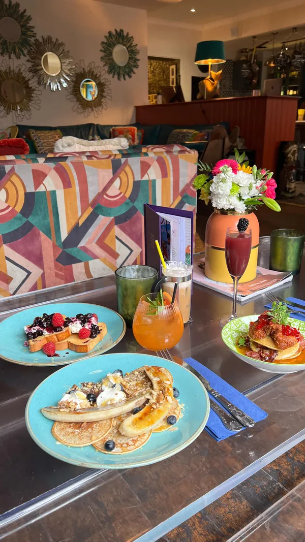 The video showcases a brunch at The Little Yellow Door in Notting Hill, London.  It features various brunch dishes, including pancakes with fried chicken and berry compote, pancakes with banana and nuts, and french toast. Cocktails and coffee drinks are also shown. The setting is a vibrant, eclectic cafe with colorful decor and plants. The presentation of the food is visually appealing, with attention to detail in plating and garnishing. No specific dietary information is provided. The main focus is on the experience of having brunch at this aesthetically pleasing cafe.