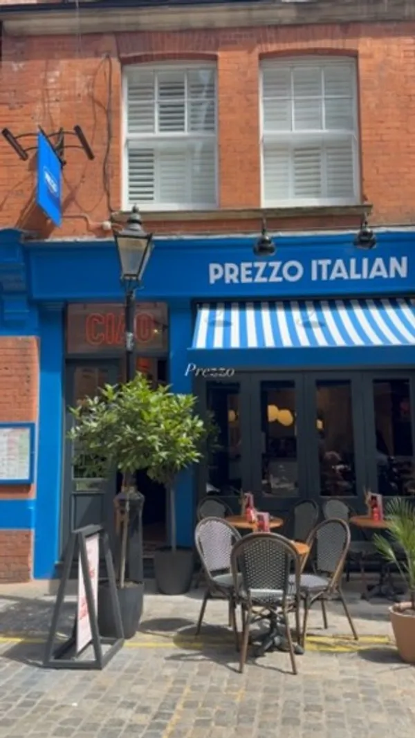 The video showcases a visit to Prezzo Italian restaurant. It begins with an exterior shot of the restaurant entrance, followed by a person entering. The interior decor, including framed artwork, is shown. Shots include a person seated at a table, the general restaurant ambiance, and close-ups of food and drinks. Specific items featured are cocktails (one purple with cherries), fried arancini balls served with tomato sauce and pesto, rigatoni pasta with a creamy sauce and grated cheese, a pizza topped with chicken, sausage, and red onion, and slices of tiramisu.
