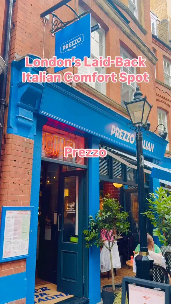 The video showcases the Prezzo Italian restaurant in Kensington, London. It begins with an exterior shot of the restaurant facade, followed by interior views of the dining area. The video features close-ups of the menu, two different drinks (a red cocktail and a yellow fizzy drink), a starter dish (oven-baked creamy mushrooms with bread), two pizzas (one pepperoni, one ham and mushroom), a side of truffle fries, and two desserts (tiramisu and Lotus Biscoff cheesecake). A hand is shown cutting a slice of the pepperoni pizza.