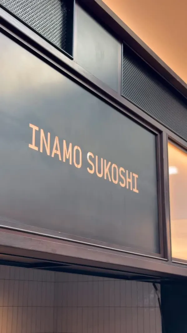 A short video montage showcasing a sushi restaurant. It begins with a shot of the restaurant's sign, 'INAMO SUKOSHI', then a digital menu board. It transitions to a wide shot of a modern food hall with a glass ceiling, followed by shots of two different sushi rolls on a wooden table. One roll is a standard uramaki roll, and the other is a creatively presented dragon roll, shaped to look like a dragon with avocado scales and carrot spikes.