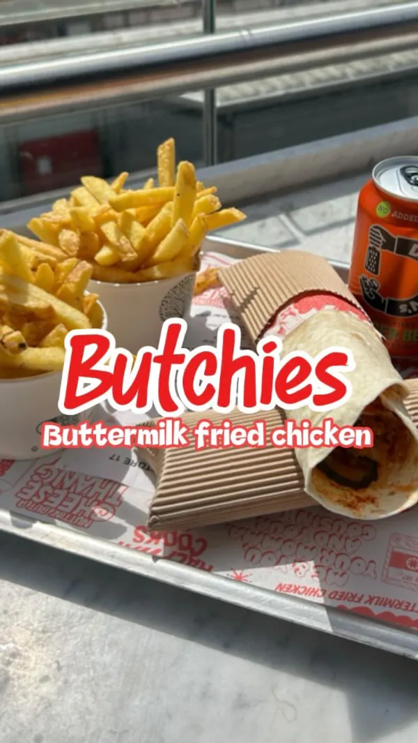 The video features a woman holding a large fried chicken wrap. It shows a tray with the wrap, fries, and drinks. It transitions to shots of the interior of Victoria Market Halls, including people eating and the Butchies Buttermilk Fried Chicken stall. The video highlights the "Butchies Bowl or Butchies Wrap with Fries or Slaw" lunch offer for £9.99. It describes the chicken as "crunchy on the outside, juicy in the inside" and "made fresh to order". The video ends with a hand taking a fry from the tray.