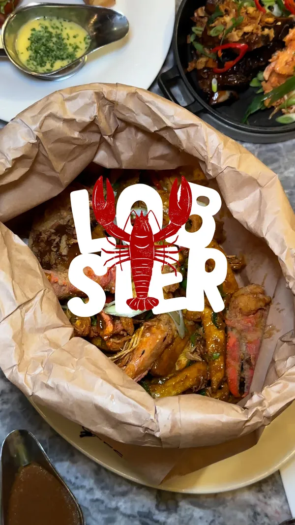 A food influencer reviews the UK's first "Lobster Spice Bag" at the restaurant Burger & Lobster. The video showcases the dish, which includes half a fried lobster, king prawns, chips, peppers, and onions in a paper bag, served with curry sauce. It features behind-the-scenes footage of the dish being prepared in the kitchen, the influencer's enthusiastic reaction while eating, and close-ups of the food's texture. Other menu items like a lobster roll, burger, ribs, and cocktails are also briefly shown.