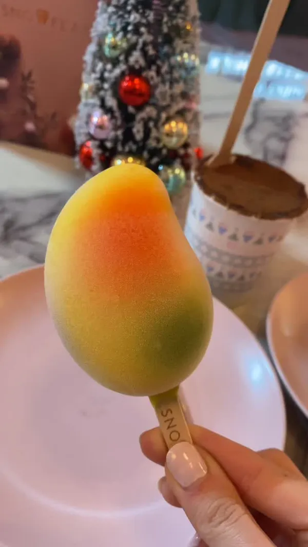 A short video showcasing the Christmas specials at Snowflake gelato. It features close-up shots of a mango-shaped gelato pop and a tiramisu cup. The video includes a shot of a display case with other festive gelato pops shaped like Santa and Christmas trees. Action shots show someone digging into the tiramisu and taking a bite of the mango gelato. The setting is a cafe with festive decorations, including a small Christmas tree on the table.