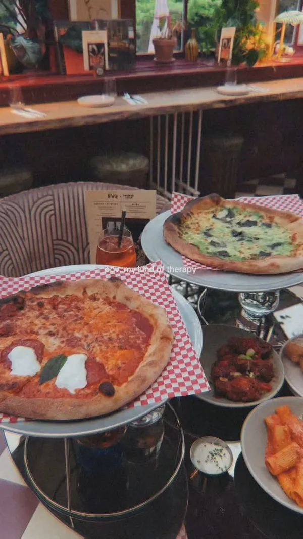 The video showcases a meal at Eve restaurant in South Kensington, London. It features various dishes including New York style pizza (half Margherita, half New Yorker), buffalo chicken wings, garlic knots with a cheese pull, rigatoni pasta with red sauce, and desserts (brownie and cheesecake). Drinks, including an Aperol Spritz and another beverage being poured, are also shown. The video includes shots of the restaurant's interior and a person eating pizza.