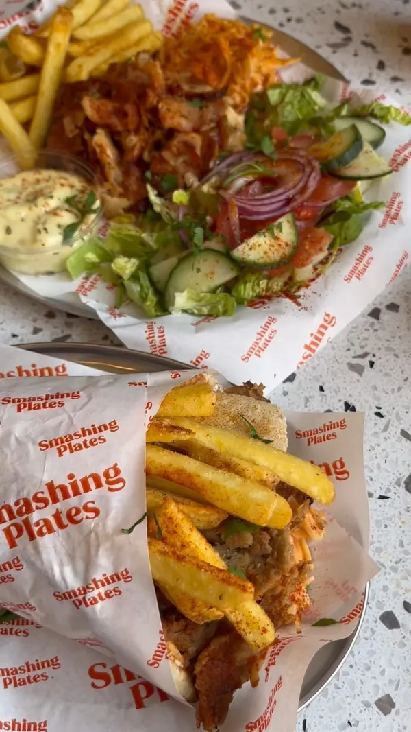 This video showcases various Greek dishes at Smashing Plates in London Bridge. The food items include chicken gyros salad, a gyro wrap with fries, halloumi fries, and baklava. The presentation is clean and appealing with consistent branding on the paper liners.  The video highlights vegetarian/vegan options with the 'Vegan Olympus Wrap'. The main focus is showcasing the food items.