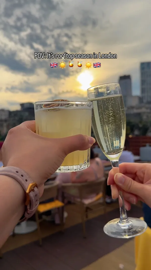 The video showcases a rooftop bar in London during rooftop season. It features various drinks including cocktails, champagne, and beer, alongside food items such as fries and small bites like sliders and canapés. The video captures people relaxing and socializing in the outdoor seating area, with views of the London skyline including landmarks like the Shard and the London Eye. The text overlay highlights that it is 'rooftop season in London'.