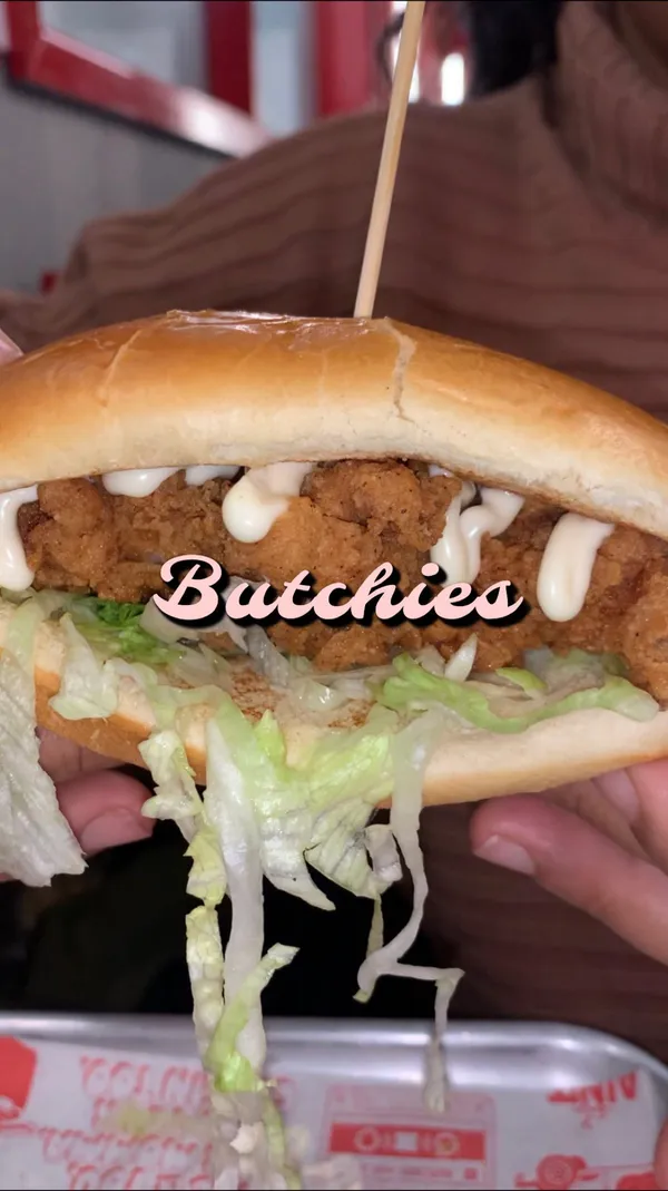 The video showcases Butchies Fried Chicken restaurant in Ealing. The main focus is on the "Chicken Dawg," a fried chicken hot dog in a brioche roll with lettuce and mayonnaise, and a chilli honey sauce.  Halloumi fries and cheesy fries are also featured. The video highlights the price (£3.95) and emphasizes the deliciousness of the food. Coca-Cola and Ting sparkling grapefruit are shown as beverage options. 