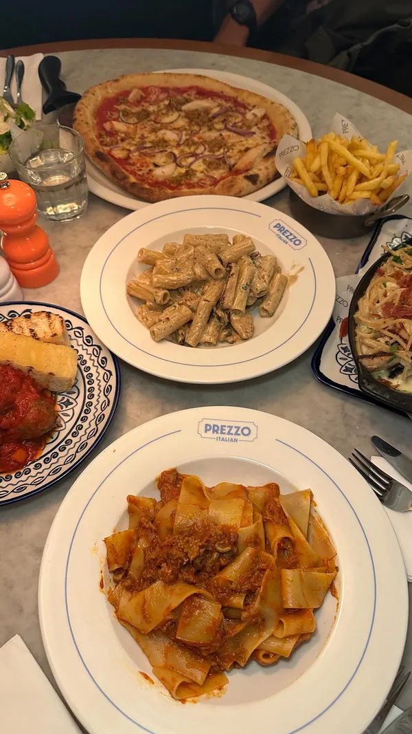 A fast-paced montage video showcasing a wide variety of Italian dishes from the restaurant Prezzo in Edinburgh. The video features numerous plates of food, including pizzas (Meat Lovers, Tuscan BBQ Chicken), pasta (Roast Duck Bolognese Pappardelle, Creamy Chicken & Pancetta Rigatoni, Crab & Lobster Ravioli, Chicken Spaghetti Carbonara Al Forno), and appetizers (Oven-baked King Prawns, Creamy Mushrooms, Pancetta Mac & Cheese Bites, Italian Meatballs). Side dishes like fries and a Caesar salad are also shown. The video includes action shots like dipping bread, grating cheese, and cutting pizza. The video concludes by highlighting desserts like Triple Cooked Nutella Doughnut Balls and Custard Cream Tiramisu.
