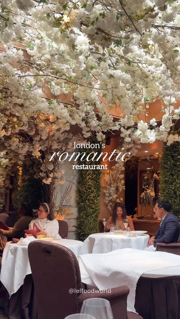 The video showcases a romantic restaurant in London's Covent Garden. It features shots of the interior with extensive white blossom decor, diners, cocktails, and various dishes including a green egg dish, stuffed eggplant, beef with sauce, salmon with vegetables, a chocolate dessert, and small pastries/chocolates. Text overlays highlight it as a romantic restaurant, mention the weekend set menu price (£37.50), and what is included (2 or 3 courses & drinks). The video concludes with an exterior shot of the restaurant and its address.