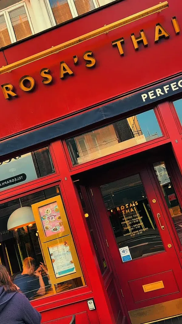The video promotes Rosa's Thai restaurant in London. It shows the exterior and interior of the restaurant, including the dining area, bar area with drinks, and wall decor. The video features various Thai dishes, including what appears to be shrimp cakes or similar fried items served with a dipping sauce, Pad Thai, and a dish with rice, minced meat (possibly basil pork or chicken), cucumber slices, and a fried egg. It also shows mango sticky rice. The influencer is shown eating the food and drinking. The presentation of the food is typical for a casual Thai restaurant. No specific dietary information is highlighted. The main focus is showcasing the food and the dining experience.