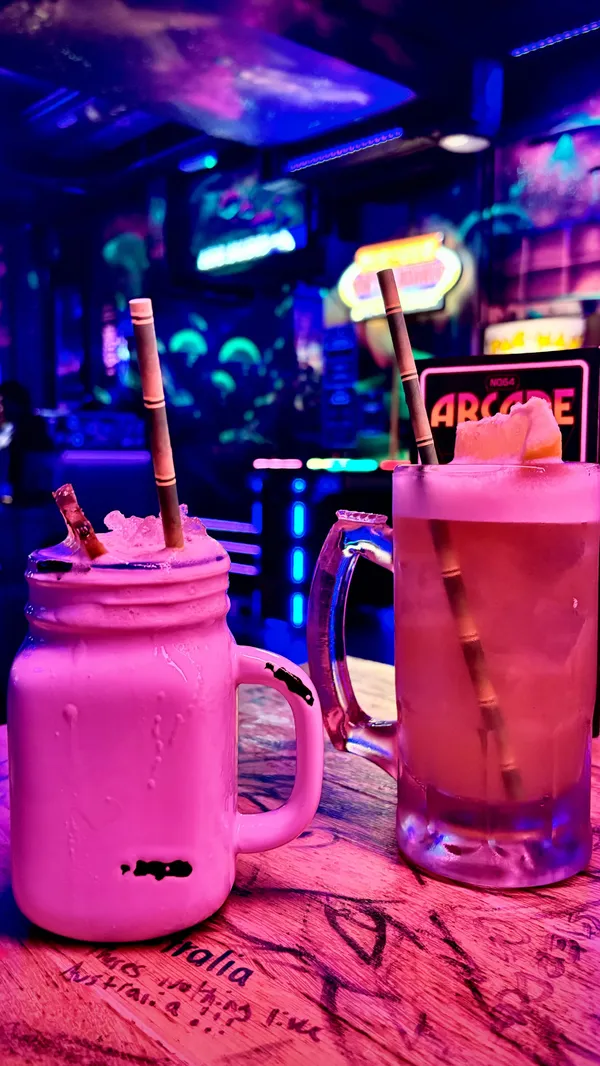 The video showcases NQ64 Arcade Bar in Newcastle. It features various arcade games like Time Crisis, Crazy Taxi, Donkey Kong, Tekken, and Dancing Stage EuroMix 2.  The bar serves game-themed cocktails; a 'Max Max zombie' cocktail is prepared and shown being flambéed.  The overall atmosphere is vibrant with neon lighting and murals. The video highlights the cost of tokens and the availability of group offers. 