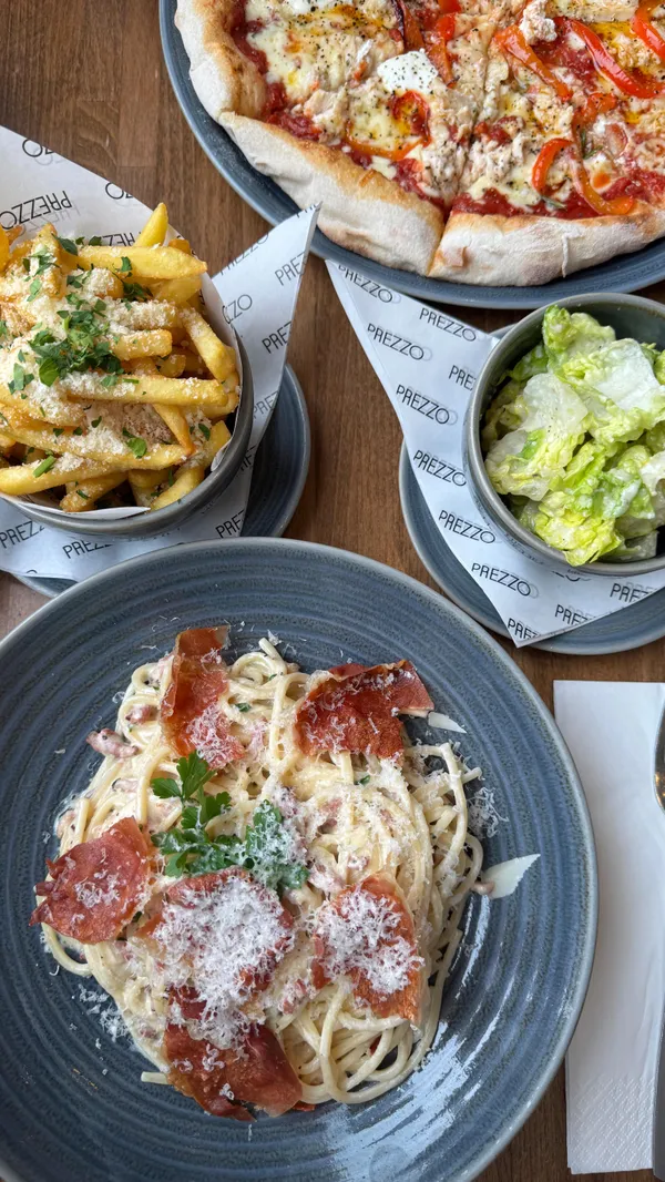 The video showcases a meal at Prezzo restaurant in Southport. It features close-ups of spaghetti carbonara being grated with cheese, truffle fries, and a chicken and roasted pepper pizza being cut and drizzled with oil. It also shows the exterior and interior of the restaurant, including the dining area and bar.