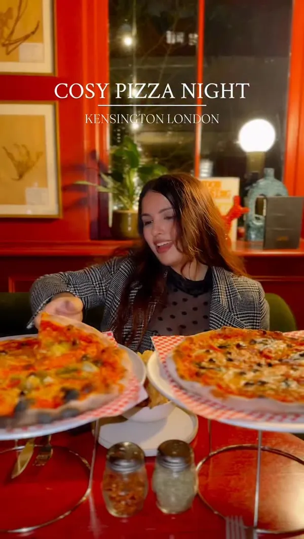 The video showcases a 'cosy pizza night' at a restaurant called Eve in Kensington, London. It features a woman enjoying two different pizzas, one of which appears to be a vegetarian option with olives, peppers, and artichokes. A side of fries and a dessert of tiramisu with pistachio gelato are also shown. The video intersperses shots of the food with clips of the restaurant's warm, dimly lit, and stylish interior, highlighting its intimate and relaxed ambiance.