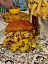 A close-up, appetizing shot of a double smash burger with melted cheese, pickles, and red onion, served on comic-book-themed paper with a side of fries in a metal cup.