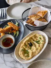 A high-quality, overhead food photography shot featuring a spread of Mediterranean-style dishes, including hummus with flatbread and halloumi fries with a dipping sauce, set on a clean marble table.