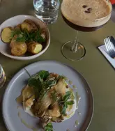 A high-quality, overhead food photography shot featuring a vegan lunch spread consisting of BBQ mushrooms with cashew cream, charred potatoes, and an espresso martini. The lighting is soft and atmospheric, typical of a modern restaurant setting, and the composition is clean and well-balanced.
