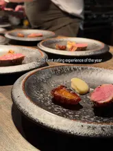 A high-quality, close-up shot of a fine-dining dish featuring a piece of seared meat, a roasted potato, and a dollop of puree, set against a blurred background of other plates.