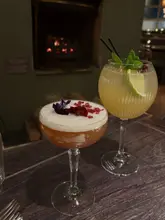 A close-up shot of two cocktails placed on a wooden table in a dimly lit pub, with a fireplace visible in the background.