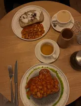 A top-down, dimly lit photograph of a brunch meal consisting of beans on sourdough toast with a green herb oil drizzle, accompanied by a coffee and a cup of tea, captured in a cafe setting.