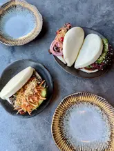 A high-quality, overhead food photograph featuring three bao buns served on textured plates against a dark, neutral background. The lighting is bright and even, highlighting the textures of the buns and the fillings.