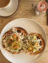 A top-down, well-composed shot of a brunch dish featuring two flatbreads topped with fresh slaw, salsa, and a drizzle of sauce, served alongside a coffee. The natural lighting and rustic wooden table setting create an inviting, casual atmosphere typical of a brunch cafe.